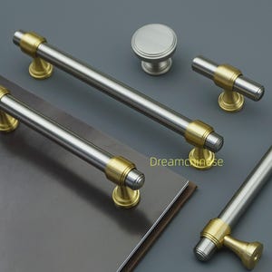 May include: A selection of cabinet handles and a knob, featuring a brushed nickel finish with gold-coloured accents. The set includes bar pulls and a T-shaped pull. The hardware is displayed on a dark grey surface.