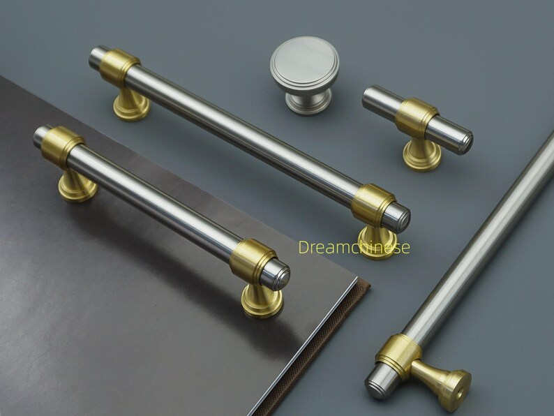 May include: A selection of cabinet handles and a knob, featuring a brushed nickel finish with gold-coloured accents. The set includes bar pulls and a T-shaped pull. The hardware is displayed on a dark grey surface.