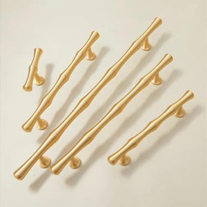 Op de afbeelding: Een verzameling goudkleurige bamboe-stijl kastgrepen en knoppen. De hardware heeft een textuur, naturalistisch ontwerp, met verschillende lengtes van grepen en een enkele knop. De hardware staat tegen een neutrale achtergrond.