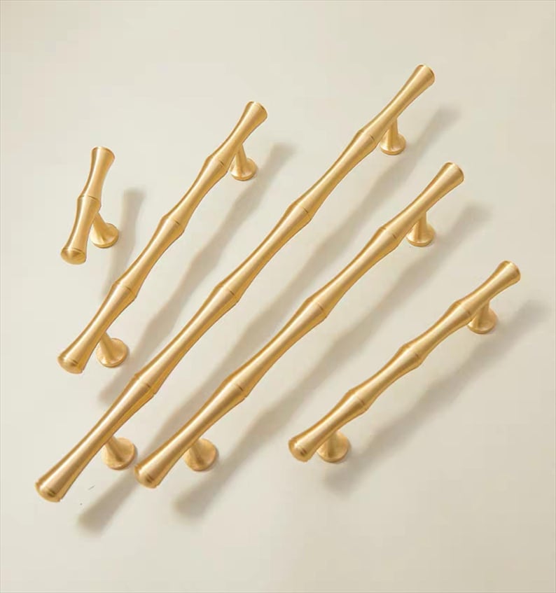 Op de afbeelding: Een verzameling goudkleurige bamboe-stijl kastgrepen en knoppen. De hardware heeft een textuur, naturalistisch ontwerp, met verschillende lengtes van grepen en een enkele knop. De hardware staat tegen een neutrale achtergrond.