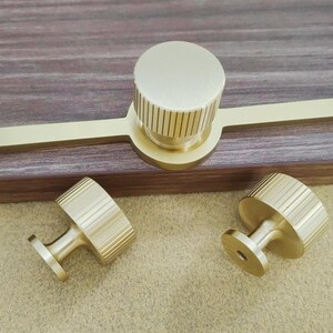 Pu&ograve; includere: Un set di tre pezzi di ferramenta per mobili in ottone. Il set comprende una lunga maniglia con un pomello circolare e due pomelli pi&ugrave; piccoli. I pomelli hanno un design a coste. L'hardware &egrave; esposto su una superficie di legno.