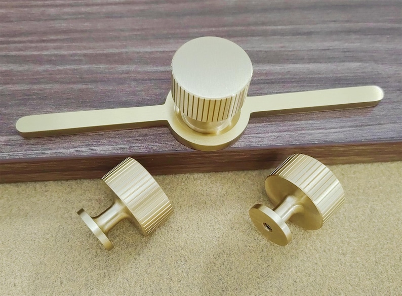 Pu&ograve; includere: Un set di tre pezzi di ferramenta per mobili in ottone. Il set comprende una lunga maniglia con un pomello circolare e due pomelli pi&ugrave; piccoli. I pomelli hanno un design a coste. L'hardware &egrave; esposto su una superficie di legno.