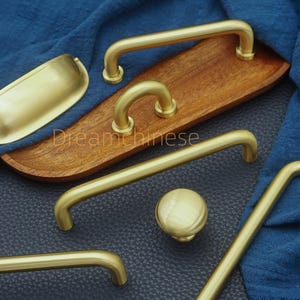 May include: A collection of brass-toned cabinet hardware, including handles, a knob, and a cup pull. The hardware is displayed on a wooden tray and a dark blue surface, with a blue fabric in the background. The Dreamchinese text is visible.