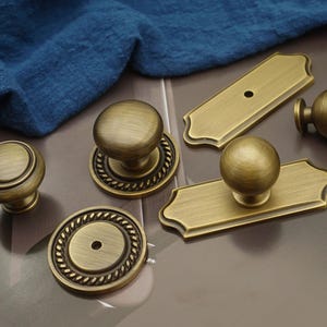 May include: An assortment of antique brass cabinet hardware, including knobs and pulls. The knobs have a round design, some with a rope-like border. The pulls are rectangular with a rounded top and a single knob. A blue cloth is in the background.