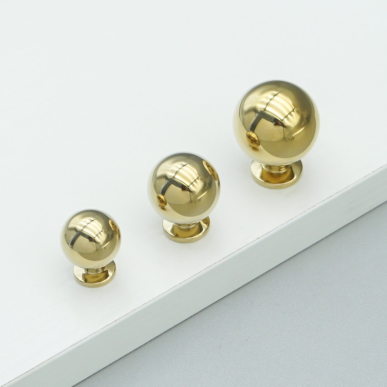 Op de afbeelding: Drie ronde gouden kastknoppen met een gladde, glanzende afwerking. De knoppen zijn van verschillende grootte, met de grootste knop aan de achterkant en de kleinste knop aan de voorkant.