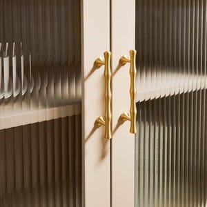 Op de afbeelding: Close-up van een kast met twee deuren. De deuren hebben geribbelde glazen panelen en zijn omlijst in een lichtbeige kleur. De kast heeft goudkleurige handgrepen in bamboestijl, wat een vleugje elegantie aan het ontwerp toevoegt.