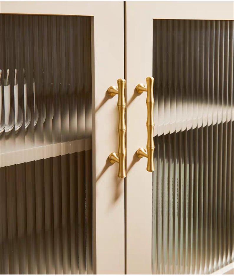 Op de afbeelding: Close-up van een kast met twee deuren. De deuren hebben geribbelde glazen panelen en zijn omlijst in een lichtbeige kleur. De kast heeft goudkleurige handgrepen in bamboestijl, wat een vleugje elegantie aan het ontwerp toevoegt.