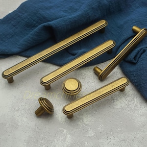 Op de afbeelding: Een verzameling antieke messing kastbeslagen, waaronder handgrepen en een knop. De handgrepen hebben een rechthoekige vorm met drie parallelle groeven. De hardware wordt gepresenteerd op een blauwe doek en een lichtgrijze ondergrond.