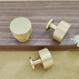 Pu&ograve; includere: Tre pomelli dorati per mobili con un design a coste. Un pomello &egrave; rotondo, mentre gli altri due hanno una forma rettangolare. I pomelli sono esposti su una superficie di legno.