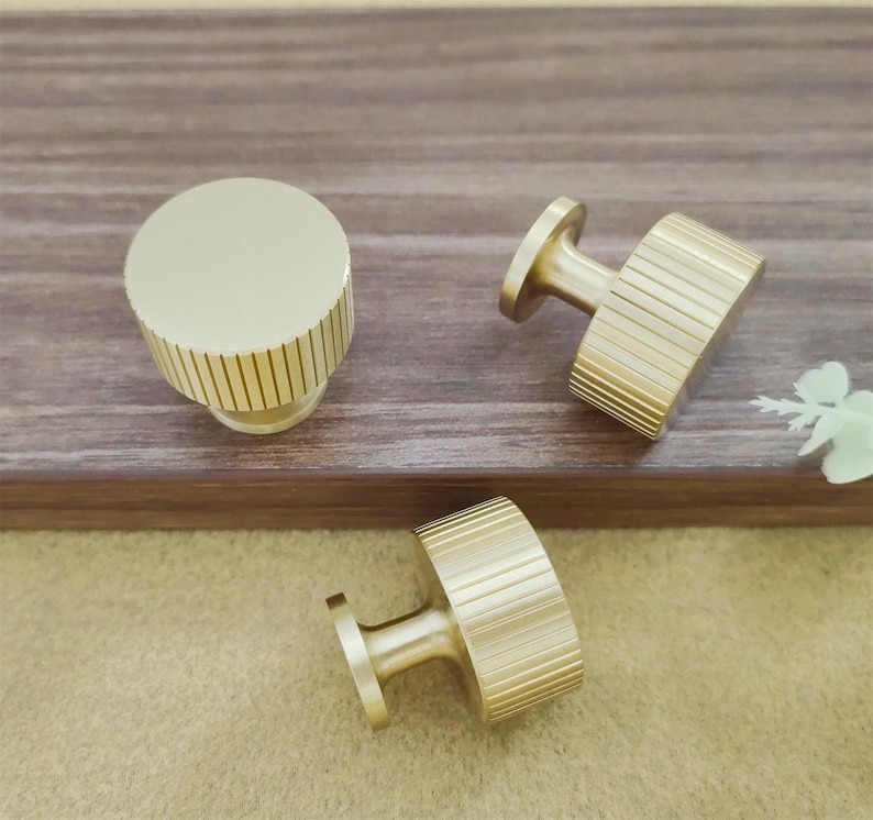 Pu&ograve; includere: Tre pomelli dorati per mobili con un design a coste. Un pomello &egrave; rotondo, mentre gli altri due hanno una forma rettangolare. I pomelli sono esposti su una superficie di legno.