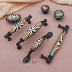 以下が含まれることがあります： 装飾的なキャビネットハードウェアのコレクション。ノブとプルが含まれています。 白、オレンジ、ターコイズの幾何学的なスターバーストデザインの黒いベースが特徴です。 Dreamchineのロゴが見えます。