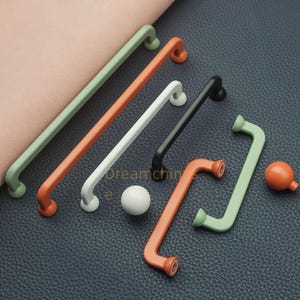 Könnte beinhalten: Eine Sammlung farbenfroher Schrankgriffe und -knöpfe in verschiedenen Formen und Größen. Die Griffe sind in Grün, Orange, Weiß und Schwarz gehalten. Eine weiße Kugel und ein oranger Knopf sind ebenfalls vorhanden. Die Hardware befindet sich vor einem strukturierten Hintergrund.