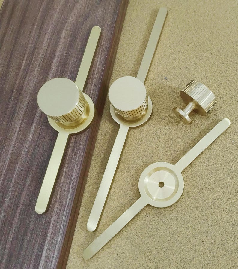 Pu&ograve; includere: Una collezione di componenti hardware in ottone, tra cui pomelli e maniglie. I pomelli hanno una forma cilindrica con dettagli scanalati, mentre le maniglie presentano un design sottile e allungato. L'hardware &egrave; esposto su una superficie di legno e una superficie beige.