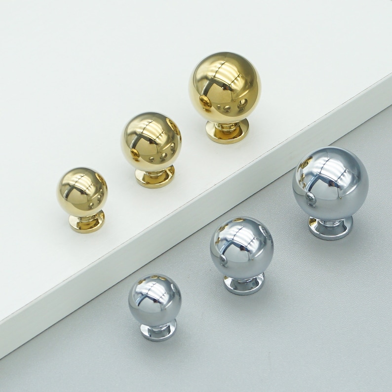 Op de afbeelding: Vijf ronde kastknoppen, twee goud en drie zilver. De knoppen zijn allemaal even groot en hebben dezelfde vorm, maar hebben verschillende afwerkingen.