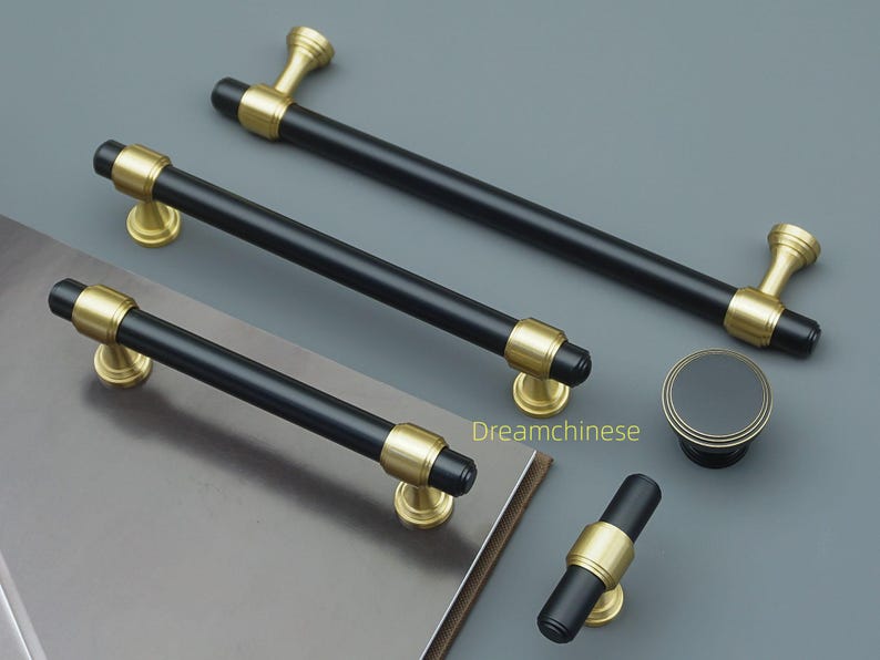 May include: A selection of black and gold cabinet hardware, including handles and a knob. The handles have a cylindrical black centre with gold accents and mounting bases. The knob is round with a black centre and gold trim. The hardware is displayed on a grey surface.