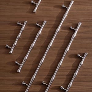 Op de afbeelding: Een verzameling zilverkleurige bamboe-stijl kastgrepen en een enkele knop, gerangschikt op een houten oppervlak. De grepen vari&euml;ren in lengte, met een centraal bamboe-achtig ontwerp en T-vormige uiteinden voor montage. Voor kasten en lades.