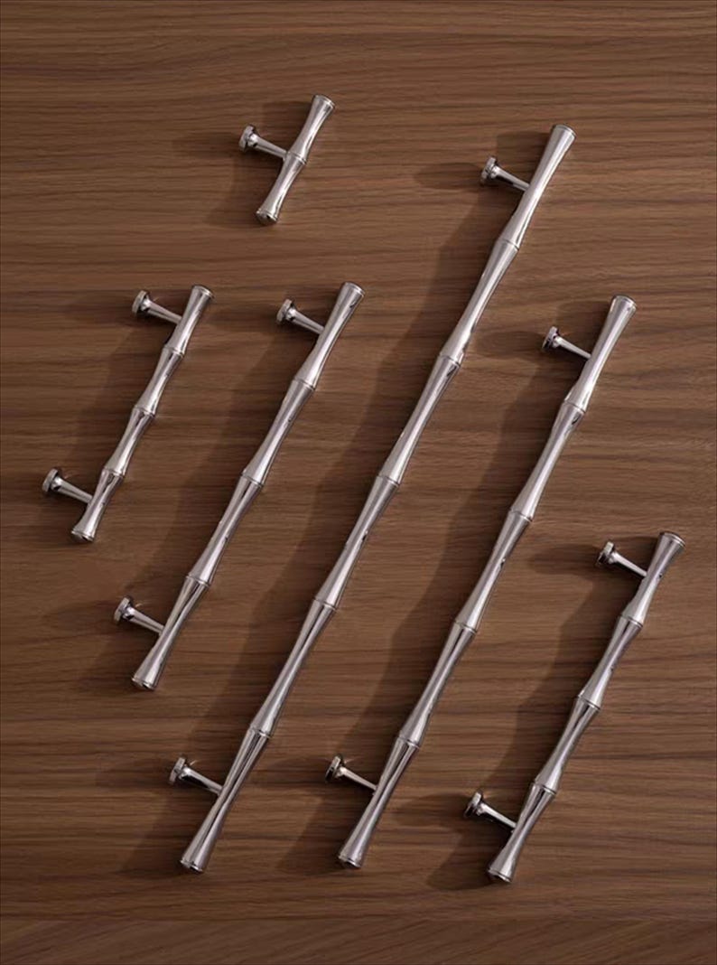 Op de afbeelding: Een verzameling zilverkleurige bamboe-stijl kastgrepen en een enkele knop, gerangschikt op een houten oppervlak. De grepen vari&euml;ren in lengte, met een centraal bamboe-achtig ontwerp en T-vormige uiteinden voor montage. Voor kasten en lades.