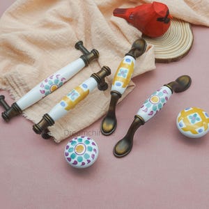 May include: A collection of decorative cabinet hardware, including handles and knobs. The handles have white ceramic centers with colorful floral patterns and bronze-toned metal accents. The knobs feature matching designs. A wooden bird and a wood slice are in the background.