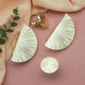 May include: Gold-colored cabinet hardware set, including two half-circle handles, a round knob, and a pull. The hardware features a sunburst design and is displayed on a pink surface with a beige cloth and greenery.