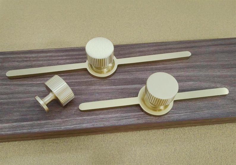 Pu&ograve; includere: Un set di ferramenta dorata per armadi, tra cui due maniglie e un pomello, disposti su una superficie in legno scuro. Le maniglie hanno un'impugnatura circolare con scanalature verticali e una barra piatta. Il pomello ha un design simile.