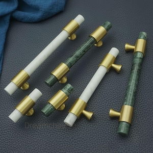 May include: A selection of cabinet handles with a contemporary design. The handles have cylindrical bars in white and green marble, complemented by gold-coloured metal fixtures. These handles are designed for simple fitting and add a touch of sophistication to any cabinet or drawer.