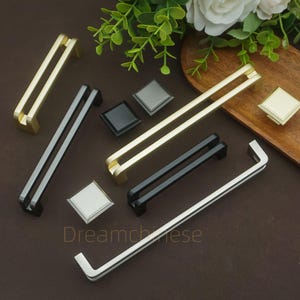 May include: An assortment of cabinet hardware, including gold, black, and silver drawer pulls and square knobs. The drawer pulls have a double bar design. The word "Dreamchiese" is printed on the surface.