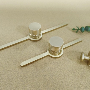 Pu&ograve; includere: Set di ferramenta per mobili color oro, tra cui due maniglie con pomolo circolare e base lineare e un pomolo abbinato. I pomoli hanno una texture a coste. L'hardware &egrave; esposto su una superficie neutra.