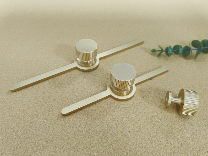 Pu&ograve; includere: Set di ferramenta per mobili color oro, tra cui due maniglie con pomolo circolare e base lineare e un pomolo abbinato. I pomoli hanno una texture a coste. L'hardware &egrave; esposto su una superficie neutra.