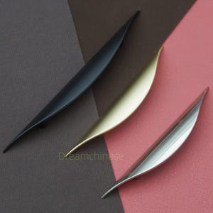 Puede incluir: Tres tiradores de armario modernos en negro, dorado y plateado. Cada tirador tiene un diseño elegante y curvado, con un extremo puntiagudo. Los tiradores están dispuestos sobre un fondo de superficies marrones y rosas. El texto "Dreamchinese" es visible.