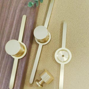 Pu&ograve; includere: Una collezione di ferramenta per mobili color oro, tra cui pomelli e maniglie. I pomelli hanno un design cilindrico con scanalature verticali, mentre le maniglie presentano una lunga e sottile impugnatura. La ferramenta &egrave; disposta su una superficie di legno e uno sfondo beige.