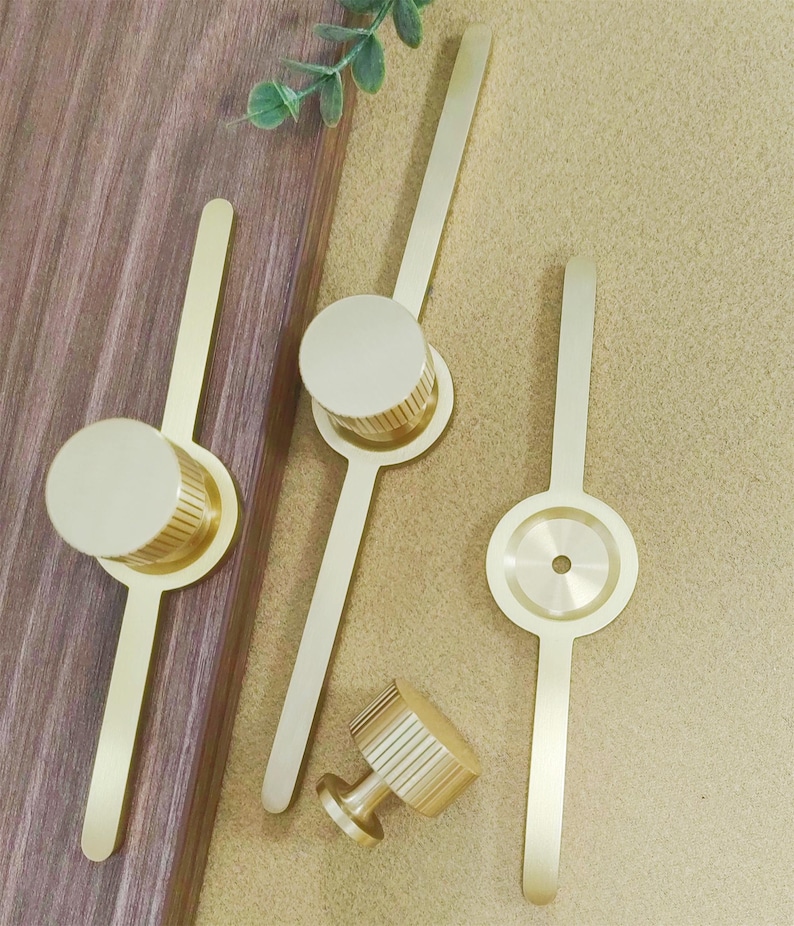 Pu&ograve; includere: Una collezione di ferramenta per mobili color oro, tra cui pomelli e maniglie. I pomelli hanno un design cilindrico con scanalature verticali, mentre le maniglie presentano una lunga e sottile impugnatura. La ferramenta &egrave; disposta su una superficie di legno e uno sfondo beige.