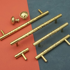 Può includere: Una collezione di ferramenta dorata per armadi, tra cui maniglie e pomelli. Le maniglie hanno un aspetto strutturato e martellato, e i pomelli hanno un design rotondo e martellato. L'hardware è esposto su uno sfondo rosso e blu.