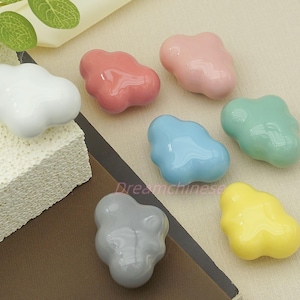 May include: Seven cloud-shaped ceramic knobs in various colours: white, red, pink, blue, green, grey, and yellow. These decorative knobs are likely for furniture or cabinets, offering a playful design element.
