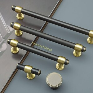 May include: A selection of black and gold cabinet handles and a knob. The bar handles have a matte black finish with gold-coloured accents. The hardware is displayed on a grey surface. The longest handle is approximately 20 cm.