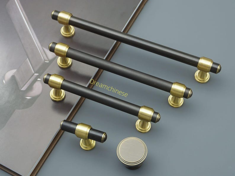 May include: A selection of black and gold cabinet handles and a knob. The bar handles have a matte black finish with gold-coloured accents. The hardware is displayed on a grey surface. The longest handle is approximately 20 cm.