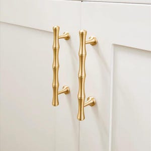 Op de afbeelding: Twee goudkleurige bamboe-stijl kastgrepen gemonteerd op een witte kastdeur. De grepen hebben een verticaal, gesegmenteerd ontwerp, dat bamboestengels nabootst, met gebogen montagebeugels.