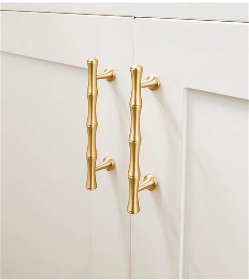Op de afbeelding: Twee goudkleurige bamboe-stijl kastgrepen gemonteerd op een witte kastdeur. De grepen hebben een verticaal, gesegmenteerd ontwerp, dat bamboestengels nabootst, met gebogen montagebeugels.