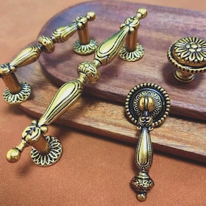 May include: An assortment of antique-style cabinet hardware in gold and black. The collection includes two pull handles, two smaller handles, and a round knob. The hardware is displayed on a wooden tray against a brown background.