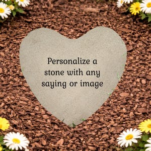 May include: A heart-shaped light gray stone with a smooth surface featuring the black printed text: Personalize a stone with any saying or image. The stone is surrounded by brown wood mulch and white and yellow daisy flowers at the corners.