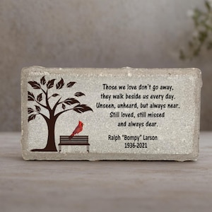 May include: A rectangular memorial stone with a textured surface. The stone features a tree, bench, and cardinal design. Text reads: "Those we love don't go away..." and includes a name and dates.