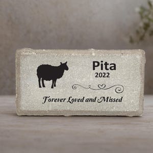 May include: A rectangular, light gray stone memorial with a black silhouette of a sheep. The name "Pita" and the year "2022" are above the text "Forever Loved and Missed". Decorative swirls and a heart accent the text.