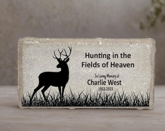 May include: A rectangular stone memorial with a black silhouette of a deer and grass. Text reads "Hunting in the Fields of Heaven" and "In Loving Memory of Charlie West 1952-2023". The stone has a textured, light gray surface.