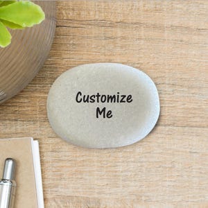 May include: A smooth, light gray oval river stone featuring the black text "Customize Me" printed in the center. The stone has a natural, rounded shape and a matte finish, providing a clean surface for personalization or decorative display.