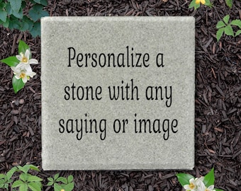 May include: A square, light gray stone paver featuring the black text "Personalize a stone with any saying or image" centered on its surface. The stone has a textured, natural finish and is positioned flat with clean, straight edges.