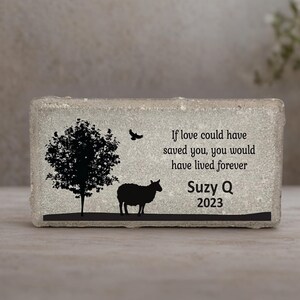 May include: A rectangular, gray stone block with a black silhouette of a tree, sheep, and bird. The text reads, "If love could have saved you, you would have lived forever. Suzy Q 2023."