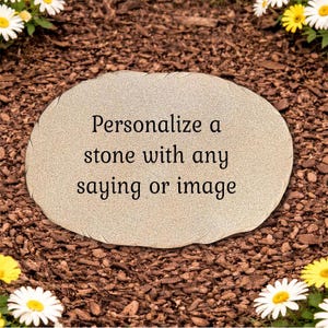 May include: An oval-shaped light beige stone with a textured surface featuring the black printed text: Personalize a stone with any saying or image. The stone has a natural, irregular edge and is positioned flat on a bed of dark brown wood mulch.