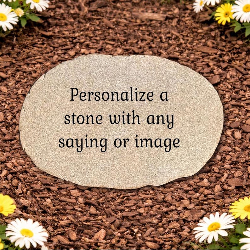 May include: An oval-shaped light beige stone with a textured surface featuring the black printed text: Personalize a stone with any saying or image. The stone has a natural, irregular edge and is positioned flat on a bed of dark brown wood mulch.