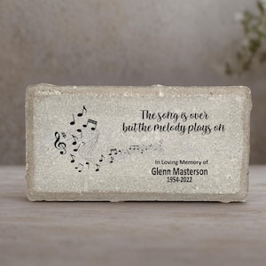 May include: A rectangular, light-colored memorial stone with the text "The song is over but the melody plays on." Black musical notes and a treble clef are on the left. The text "In Loving Memory of Glenn Masterson 1954-2022" is below.