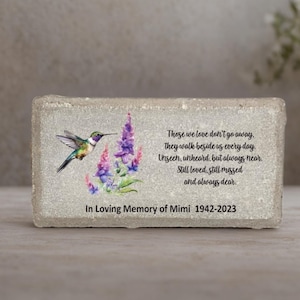 May include: A rectangular memorial stone with a hummingbird and floral design. The hummingbird is depicted with green, blue, and yellow feathers. Text reads "Those we love don't go away... In Loving Memory of Mimi 1942-2023."