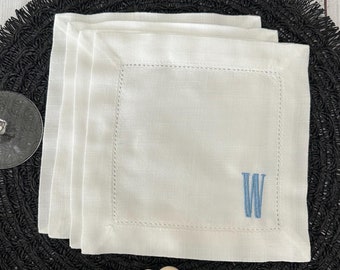 May include: A stack of square white linen napkins featuring a hemstitched border and a light blue embroidered letter W in the bottom right corner. The napkins are layered neatly, showcasing the fabric texture and the precise stitching detail.